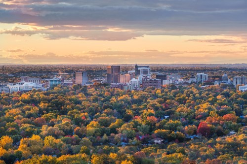 A panoramic view of a city skyline surrounded by vibrant autumn foliage under a colorful sunset.