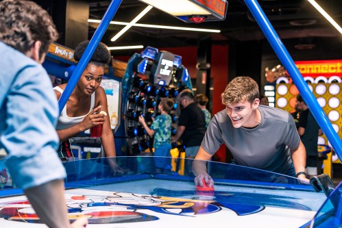 Two young adults play air hockey in a vibrant arcade, surrounded by various games and other players.