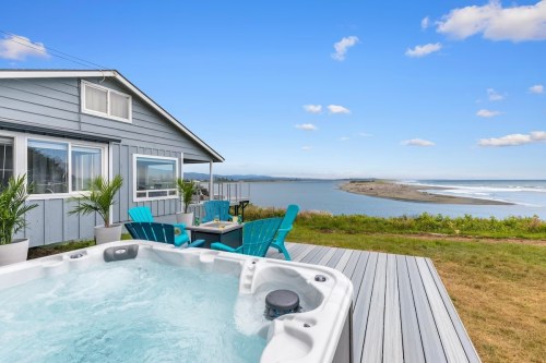 A hot tub on a deck overlooks a scenic view of a river and coastline, with a blue house in the background.