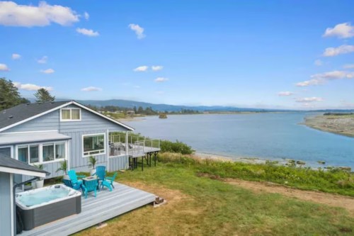 A coastal home with a hot tub and blue chairs, overlooking a serene waterway and distant mountains under a clear sky.