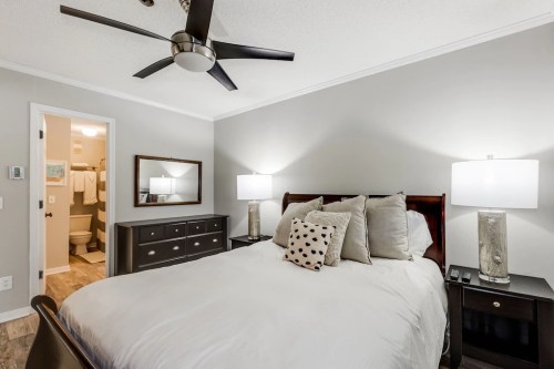 Cozy bedroom with a bed, decorative pillows, nightstands, and a bathroom visible in the background. Modern decor.