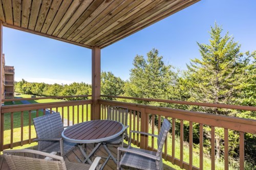 A wooden balcony with a round table and chairs, overlooking green trees and a clear blue sky.