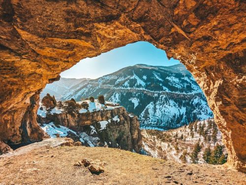 A scenic view from a cave opening, showcasing snow-covered mountains and a clear blue sky.