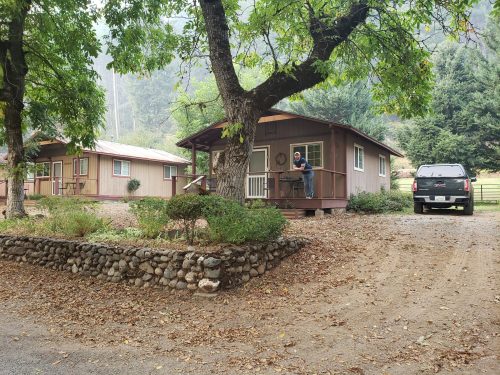 A man sits on the porch of a cabin surrounded by trees, with another cabin and a truck nearby on a gravel path.