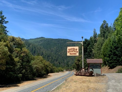 Sign for Marble Mountain Ranch along a road, surrounded by lush green trees and mountains under a clear blue sky.