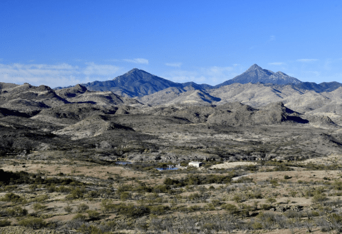 A panoramic view of rugged mountains under a clear blue sky, with sparse vegetation and a small structure in the foreground.