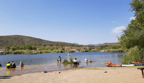 People enjoying a sunny day by a calm lake, with kayaks and lush greenery in the background.
