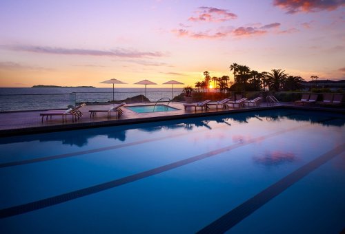 A serene poolside view at sunset, with palm trees and umbrellas reflecting in the water.