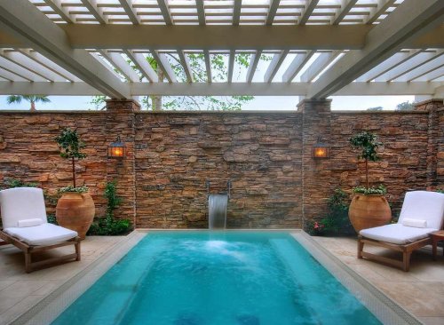 A serene spa area featuring a small pool, stone walls, and comfortable lounge chairs under a pergola.