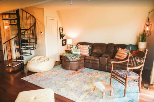 Cozy living room with a brown sofa, colorful pillows, a spiral staircase, and a bean bag chair on a patterned rug.