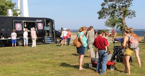 A food truck by the water with a crowd of people enjoying the outdoors on a sunny day.