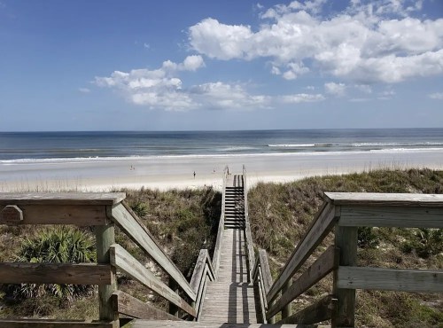 A wooden boardwalk leads to a sandy beach with gentle waves and a clear blue sky dotted with clouds.