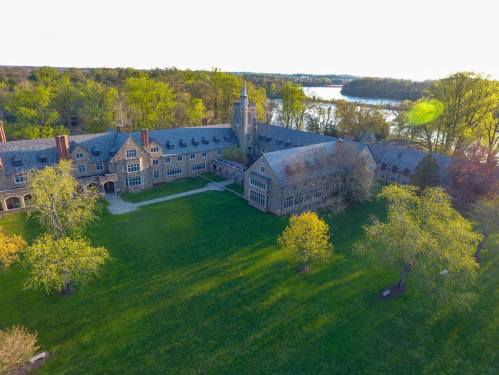 Aerial view of a large, historic stone building surrounded by lush greenery and a river in the background.