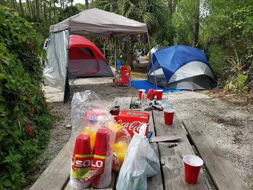 A campsite with tents, picnic tables, and scattered drinks and snacks in a wooded area.