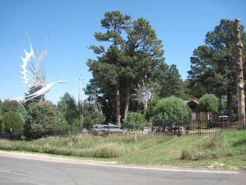 A scenic view of a street with tall trees and artistic metal sculptures in a green landscape under a clear blue sky.