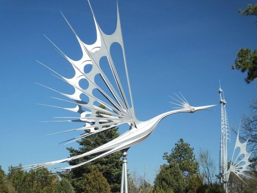 A large, metallic sculpture of a bird in flight, set against a clear blue sky and surrounded by trees.