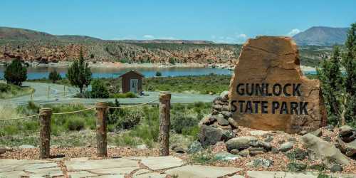 Sign marking Gunlock State Park, with a scenic view of a lake and mountains in the background. Clear blue sky above.