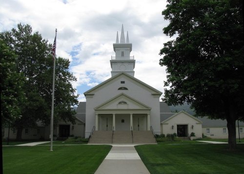 A white church building with a tall steeple, flanked by trees and a flagpole, set against a cloudy sky.