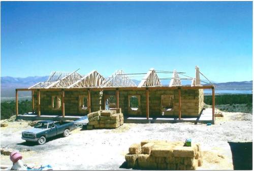 A partially constructed house made of straw bales, with wooden framing and a clear blue sky in the background.