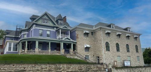 A large, colorful Victorian house next to a stone building under a blue sky with scattered clouds.