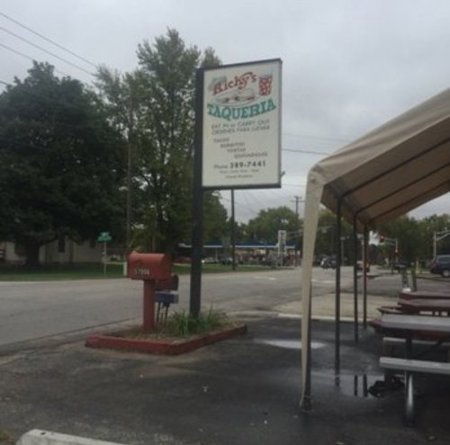 Sign for Ricky's Taqueria, with outdoor seating and a mailbox nearby, set on a cloudy day.