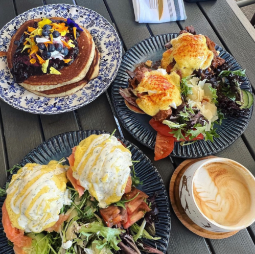 A table with three plates: pancakes with berries, eggs Benedict with salad, and a latte in a wooden cup.