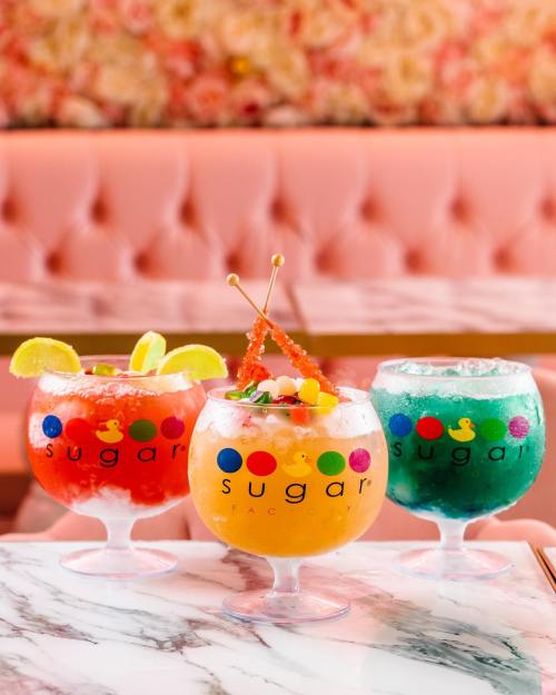 Three colorful cocktails in large glasses, featuring vibrant garnishes, set against a pink floral backdrop.