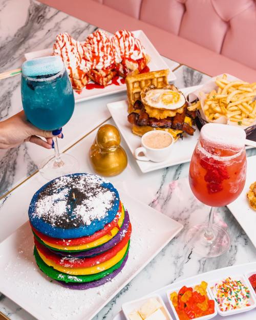 A colorful stack of rainbow pancakes with drinks and various dishes on a marble table, featuring a rubber duck decoration.
