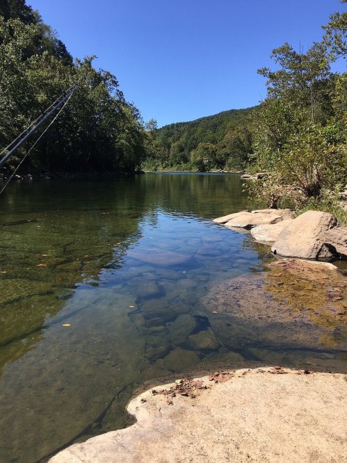 A serene river scene with clear water, rocky banks, and lush greenery under a bright blue sky.