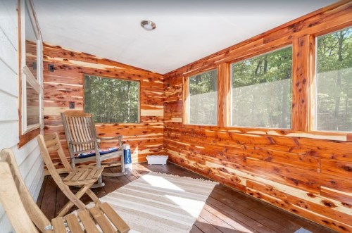 Cozy sunroom with wooden walls, two chairs, and a light rug, surrounded by large windows.