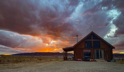 A rustic cabin against a dramatic sunset sky with colorful clouds and distant mountains.