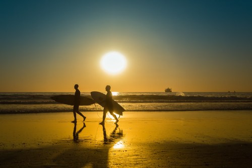 Two surfers walk along a beach at sunset, with the sun setting over the ocean in the background.