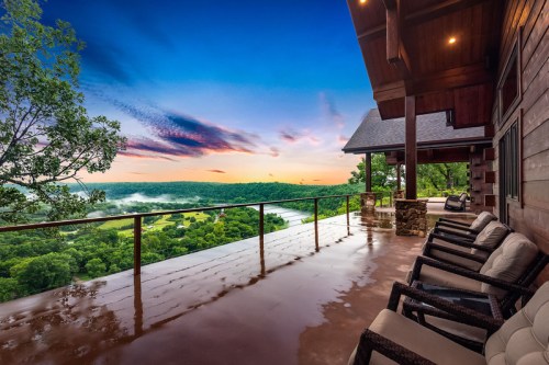A scenic view from a covered porch overlooking a lush valley at sunrise, with comfortable seating and a clear sky.