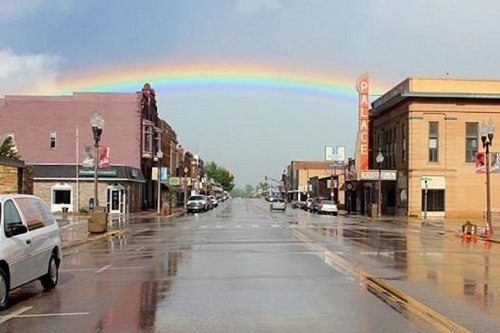 A vibrant rainbow arches over a wet street lined with historic buildings and parked cars after a rain shower.