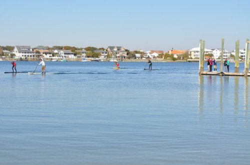 People paddleboarding on a calm lake with houses in the background and a dock on the right. Clear blue sky.