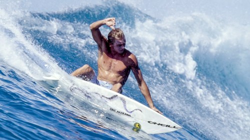 A shirtless surfer skillfully rides a wave on his surfboard, showcasing dynamic movement and ocean spray.