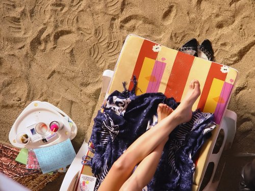 Aerial view of a beach chair with a person’s legs resting on it, surrounded by sand and personal items.