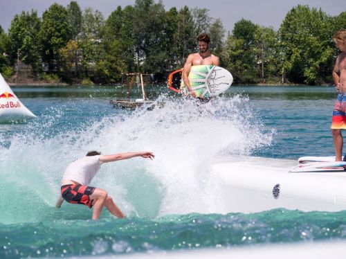 A surfer falls into the water while another prepares to ride a wave, with trees and a boat in the background.
