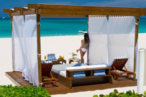 A beach cabana with white curtains, a sunbed, and a woman in a bikini overlooking the turquoise ocean.