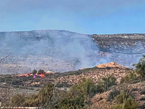 Smoke rises from a hillside with emergency vehicles visible in the distance, indicating a fire in the area.