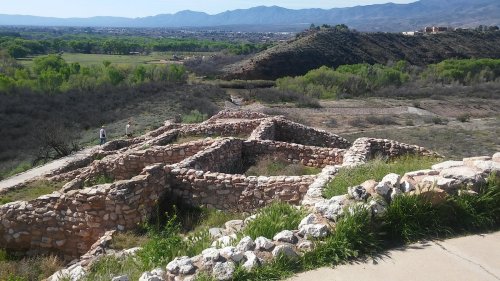 Ancient stone ruins on a hillside, surrounded by greenery and mountains in the background under a clear blue sky.
