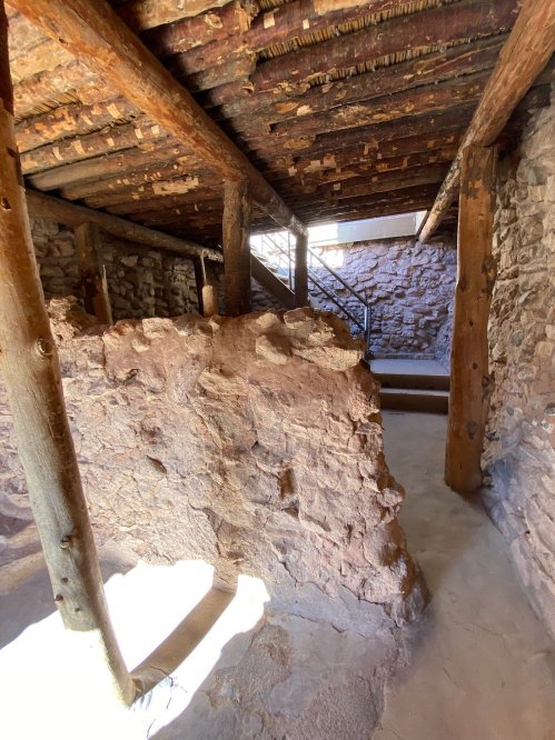 Interior view of a rustic stone structure with wooden beams and a staircase, illuminated by natural light.