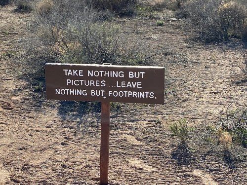 A wooden sign in a desert area reads: "Take nothing but pictures... leave nothing but footprints."