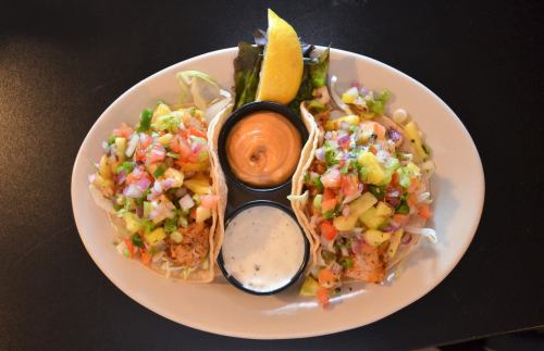 Two tacos filled with colorful toppings, served with dipping sauces and a lemon wedge on a white plate.