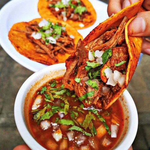 A hand holds a taco filled with meat, dipping it into a bowl of broth, with more tacos in the background.