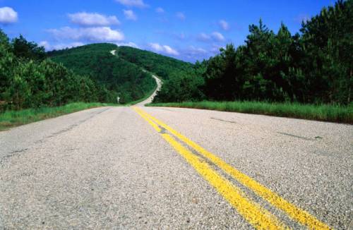 A winding road stretches through green hills under a blue sky with fluffy clouds.
