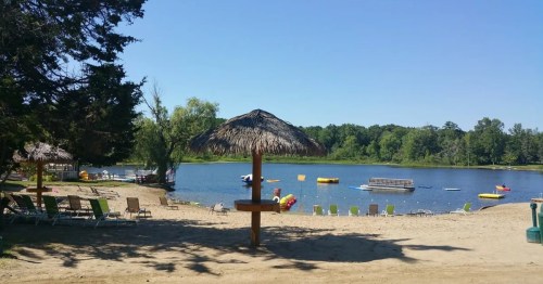 A sandy beach with a thatched umbrella, lounge chairs, and a calm lake featuring colorful inflatable toys.