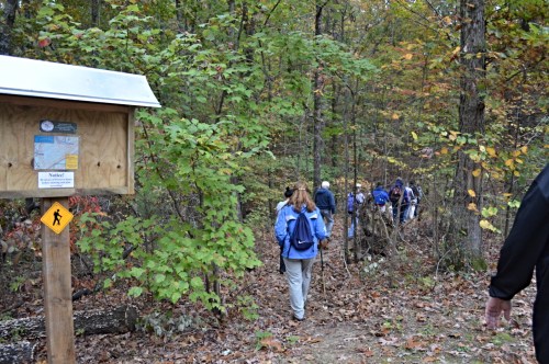 A group of hikers walks along a wooded trail, with a trail sign and autumn foliage surrounding them.