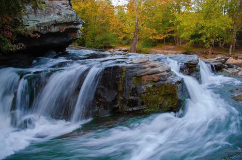 A serene waterfall cascades over rocky ledges, surrounded by vibrant autumn foliage and a tranquil stream.