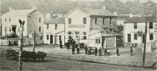 Historic street scene featuring a telegraph office, buildings, and people gathered outside, with horses nearby.